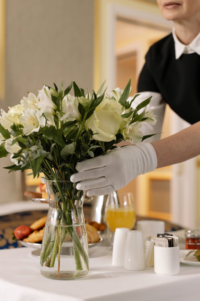 services-01 Woman arranging flowers in a hotel room with breakfast on a table, emphasizing luxury and convenience.