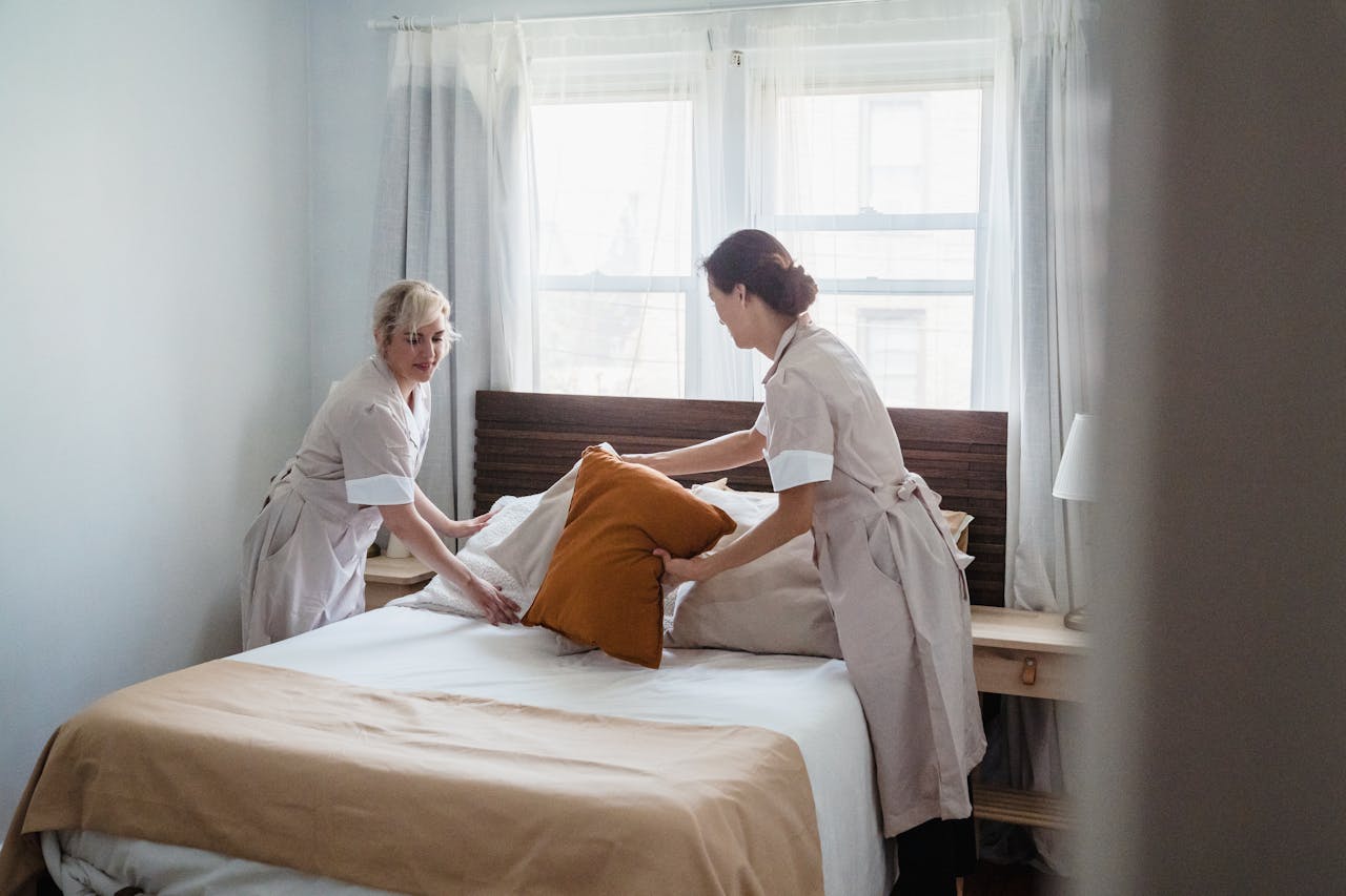 about-img Two housekeepers organizing bed linen in a bright, cozy bedroom.